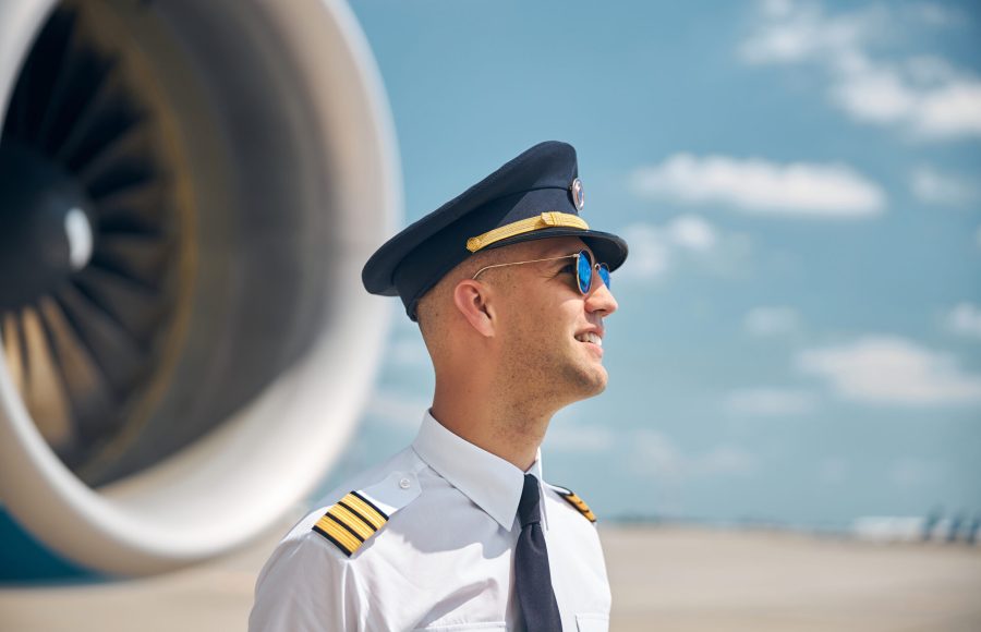 Handsome young man in captain hat looking away and smiling while standing at airport with airplane and sky on blurred background